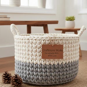 Pode incluir: Cesto de crochê feito à mão, de duas cores, com secções creme e cinza. O cesto tem duas alças e uma etiqueta castanha que diz "Craftlooms Works - Handmade With Love". Dois pinhas estão em frente ao cesto.