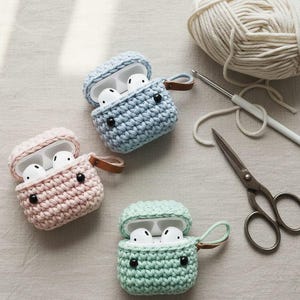Può includere: Tre custodie per AirPod all'uncinetto in colori pastello: rosa, blu e verde. Ogni custodia ha un passante in pelle e occhi di bottone neri. Gli AirPods sono bianchi. Un gomitolo di lana, un uncinetto e delle forbici sono anche nell'immagine.