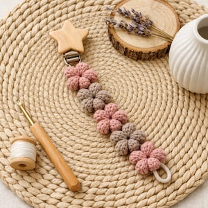 May include: A handmade baby pacifier clip with a wooden star and a series of crocheted flowers in pink and taupe. The clip is attached to a metal clasp. A crochet hook and spool of thread are also visible.