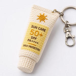 May include: A crocheted keychain shaped like a sunscreen tube. The tube is off-white with a yellow label that reads "SUN CARE 50+ SPF PA++++ DAILY PROTECTION". It has a silver key ring and clasp.