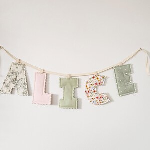Puede incluir: Una guirnalda de letras de tela que forma el nombre ALICE, con letras acolchadas individuales en varios patrones y colores, incluyendo motivos florales, rosa liso, verde salvia liso y lunares blancos, suspendidas de una cinta color crema.