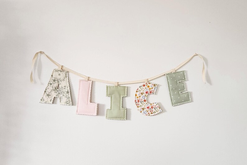 Puede incluir: Una guirnalda de letras de tela que forma el nombre ALICE, con letras acolchadas individuales en varios patrones y colores, incluyendo motivos florales, rosa liso, verde salvia liso y lunares blancos, suspendidas de una cinta color crema.