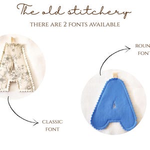 Puede incluir: Dos adornos de letra A de tela con un lazo para colgar. La letra izquierda tiene un patr&oacute;n floral beige etiquetado como CLASSIC FONT. La letra derecha es azul s&oacute;lido y est&aacute; etiquetada como ROUND FONT. El texto superior dice The old stitchery THERE ARE 2 FONTS AVAILABLE.