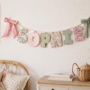 Puede incluir: Un cartel de letras de tela que deletrea el nombre SOPHIE, con letras individuales en varios patrones florales y colores pastel s&oacute;lidos. El cartel est&aacute; suspendido por un cord&oacute;n claro y decorado con un lazo rosa a la izquierda y uno verde a la derecha.