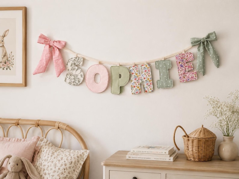 Puede incluir: Un cartel de letras de tela que deletrea el nombre SOPHIE, con letras individuales en varios patrones florales y colores pastel s&oacute;lidos. El cartel est&aacute; suspendido por un cord&oacute;n claro y decorado con un lazo rosa a la izquierda y uno verde a la derecha.