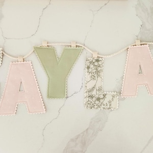 Puede incluir: Un cartel de letras de tela que deletrea el nombre ZAYLA, con letras alternas en estampado floral, verde salvia s&oacute;lido y rosa claro s&oacute;lido. Cada letra tiene un borde festoneado y est&aacute; suspendida de una cuerda trenzada de color crema.