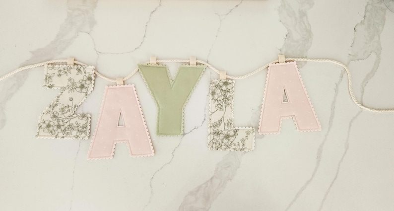 Puede incluir: Un cartel de letras de tela que deletrea el nombre ZAYLA, con letras alternas en estampado floral, verde salvia s&oacute;lido y rosa claro s&oacute;lido. Cada letra tiene un borde festoneado y est&aacute; suspendida de una cuerda trenzada de color crema.