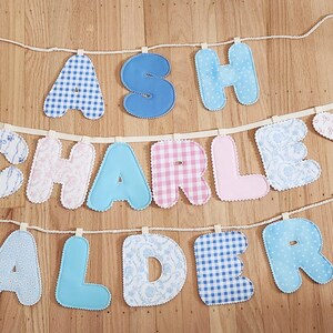 Puede incluir: Una guirnalda decorativa de tela con tres filas de letras acolchadas que forman los nombres ASH, CHARLEY y ALDER. Las letras est&aacute;n confeccionadas con diversos textiles en tonos azules, rosas y blancos, con estampados de cuadros, flores y colores lisos.