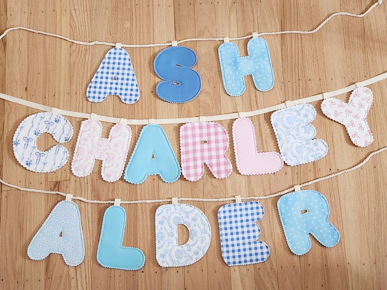 Puede incluir: Una guirnalda decorativa de tela con tres filas de letras acolchadas que forman los nombres ASH, CHARLEY y ALDER. Las letras est&aacute;n confeccionadas con diversos textiles en tonos azules, rosas y blancos, con estampados de cuadros, flores y colores lisos.