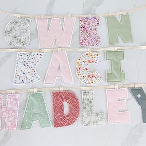 Puede incluir: Un bander&iacute;n de letras de tela con los nombres GWEN, KACI y HADLEY. Cada letra est&aacute; confeccionada con diversas telas estampadas y lisas, incluyendo motivos florales, lunares y colores s&oacute;lidos rosa, verde y gris, con bordes en blanco.