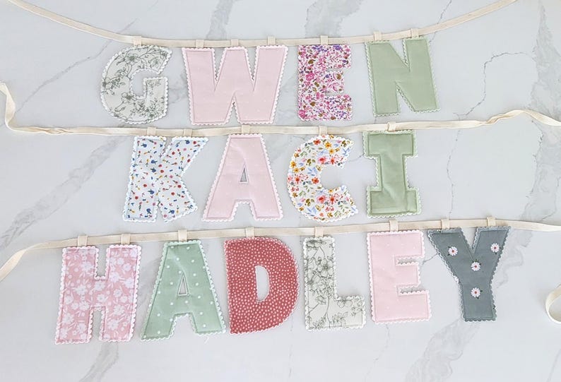 Puede incluir: Un bander&iacute;n de letras de tela con los nombres GWEN, KACI y HADLEY. Cada letra est&aacute; confeccionada con diversas telas estampadas y lisas, incluyendo motivos florales, lunares y colores s&oacute;lidos rosa, verde y gris, con bordes en blanco.