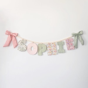 Puede incluir: Una pancarta de tela que deletrea el nombre SOPHIE con letras individuales unidas a una cuerda. Las letras presentan varios patrones florales y pasteles s&oacute;lidos en rosa, verde y blanco. Un lazo rosa est&aacute; a la izquierda y uno verde a la derecha.