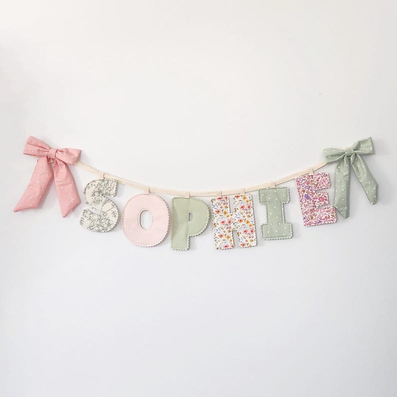 Puede incluir: Una pancarta de tela que deletrea el nombre SOPHIE con letras individuales unidas a una cuerda. Las letras presentan varios patrones florales y pasteles s&oacute;lidos en rosa, verde y blanco. Un lazo rosa est&aacute; a la izquierda y uno verde a la derecha.