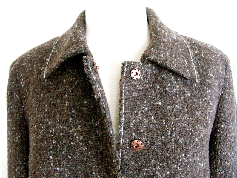 May include: A brown tweed jacket with a collar and two buttons. The jacket has a speckled pattern and a white stitching detail.