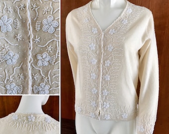 Puede incluir: Un cárdigan de color crema con bordados de flores y enredaderas en cuentas blancas. El cárdigan tiene cuello en V y mangas largas.