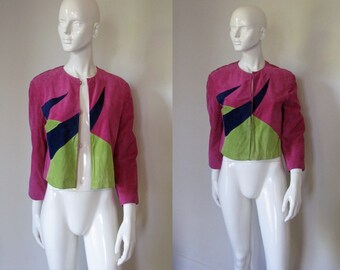 May include: A pink suede jacket with geometric green and blue accents. The jacket has a zipper closure and long sleeves.