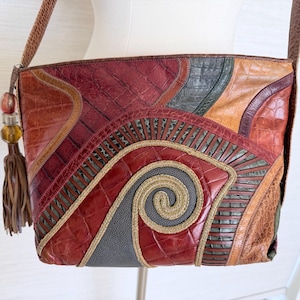 May include: A shoulder bag featuring a patchwork design of red, brown, and gray textured leather with a crocodile-skin pattern. The front displays a central spiral motif outlined in gold-toned cord, accented by a brown leather tassel with beads.