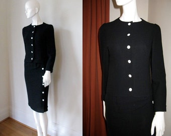 May include: A black skirt suit with a white button closure. The jacket has long sleeves and a round neckline. The skirt is knee-length and has a straight silhouette.