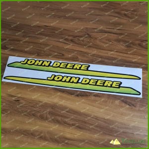May include: Two John Deere decals. Each decal has the words "JOHN DEERE" in bold, yellow letters with a black outline, set against a green and yellow gradient background. The decals are on a white background.