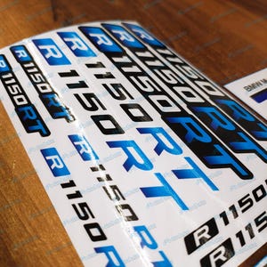 Puede incluir: Una hoja de pegatinas blancas con el texto "R 1150 RT" en negro y azul. Las pegatinas son para una motocicleta BMW. El texto está en negrita, fuente sans-serif. Las pegatinas están dispuestas en filas.