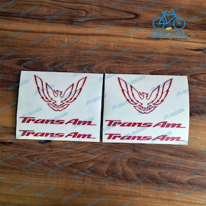 May include: Two white decals with a red graphic of a bird and the text "Trans Am". The decals are on a wooden surface. The text "AlexCycles" is printed on the decals.
