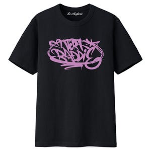 Könnte beinhalten: Schwarzes T-Shirt mit Rundhalsausschnitt und kurzen Ärmeln. Das Shirt zeigt den Text "VERA BADDIE" in einem hellvioletten Graffiti-Stil. Der Markenname "La Maglieria" ist auf dem Kragen aufgedruckt.