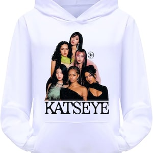 May include: White hoodie with a graphic of six women. The word "KATSEYE" is printed in black block letters at the bottom of the image. The hoodie has a drawstring and a front pocket.