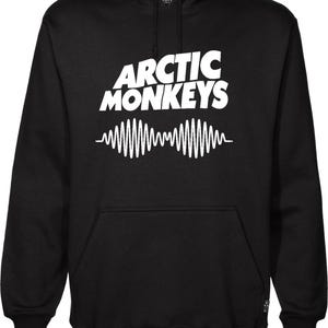 Pode incluir: Moletom preto com o nome da banda "Arctic Monkeys" em letras brancas e um design de onda sonora branco. O moletom tem um bolso frontal e um capuz com cordão. Os punhos e a bainha são canelados.