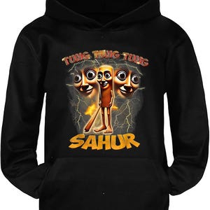 Op de afbeelding: Zwarte hoodie met een cartoonontwerp. De afbeelding toont een personage met drie gezichten, bliksem en het woord "SAHUR" in geel. De tekst "TUNG TUNG TUNG" is in rood en geel.