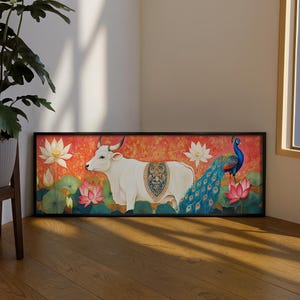 Könnte beinhalten: Ein gerahmtes Kunstwerk mit einem weißen Bullen mit dekorativem Sattel, einem Pfau und Lotusblumen vor rotem Hintergrund. Das Kunstwerk befindet sich in einem schwarzen Rahmen und liegt auf einem Holzboden.