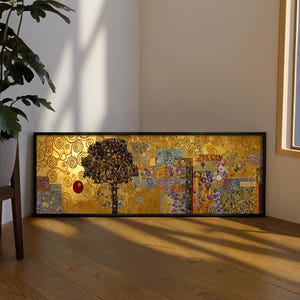 Può includere: Una stampa artistica incorniciata raffigurante un albero stilizzato ed elementi floreali su sfondo dorato. L'opera d'arte ha una cornice nera ed è esposta su un pavimento in legno. Lo stile ricorda l'Art Nouveau, con una sfera rossa.