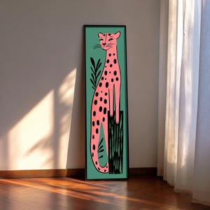 May include: A framed art print featuring a pink cheetah with black spots against a teal background. The cheetah is depicted in a stylized, seated pose. Black lines represent foliage and a base. The frame is black.