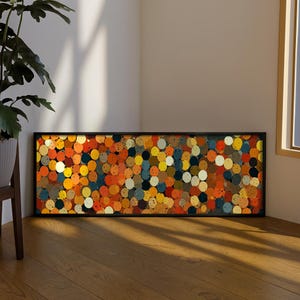 Puede incluir: Una obra de arte rectangular enmarcada con un diseño abstracto de círculos de colores. Los círculos son en tonos de naranja, amarillo, azul y marrón. El marco es negro y la obra se exhibe sobre un suelo de madera.