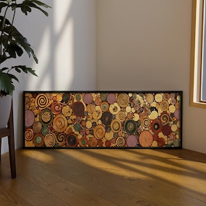 Peut inclure: Une œuvre d'art abstraite encadrée, présentant une mosaïque de cercles tourbillonnants colorés dans des tons or, marron, rose et vert. L'œuvre est posée sur un fond doré et encadrée en noir, créant un contraste visuel saisissant. L'ensemble est posé sur un sol en bois.