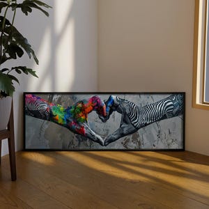 Op de afbeelding: Een rechthoekig ingelijst kunstwerk aan de muur met een grafische illustratie van twee handen die een hartvorm vormen. De ene hand is versierd met levendige, veelkleurige verfspatten, terwijl de andere een zwart-wit zebrapatroon vertoont.