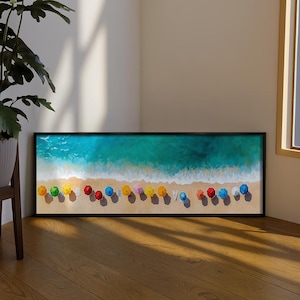 Pode incluir: Uma obra de arte emoldurada que retrata uma cena de praia com uma moldura preta. A obra mostra uma praia arenosa com guarda-sóis coloridos e água turquesa. Os guarda-sóis são de várias cores, incluindo amarelo, vermelho e azul.