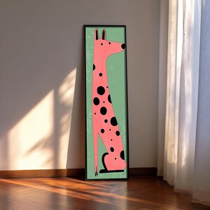 May include: A tall, rectangular framed wall art piece featuring a stylised, long-necked pink dog with black spots. The dog is depicted in a seated position against a solid light green background, enclosed within a thin, dark-coloured frame.