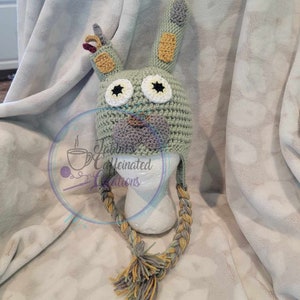 May include: A green crocheted hat with a cartoon monster face. The hat has long, braided ear-flaps with a tassel at the end.