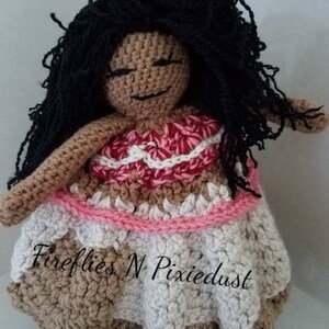 Puede incluir: Mu&ntilde;eca de crochet con piel marr&oacute;n, cabello negro y un vestido rosa y blanco. La mu&ntilde;eca lleva un vestido rosa y blanco con una falda marr&oacute;n. La mu&ntilde;eca sostiene una flor en su mano. La mu&ntilde;eca est&aacute; sentada sobre un fondo blanco. El texto "Fireflies N Pixiedust" es visible en la falda de la mu&ntilde;eca.