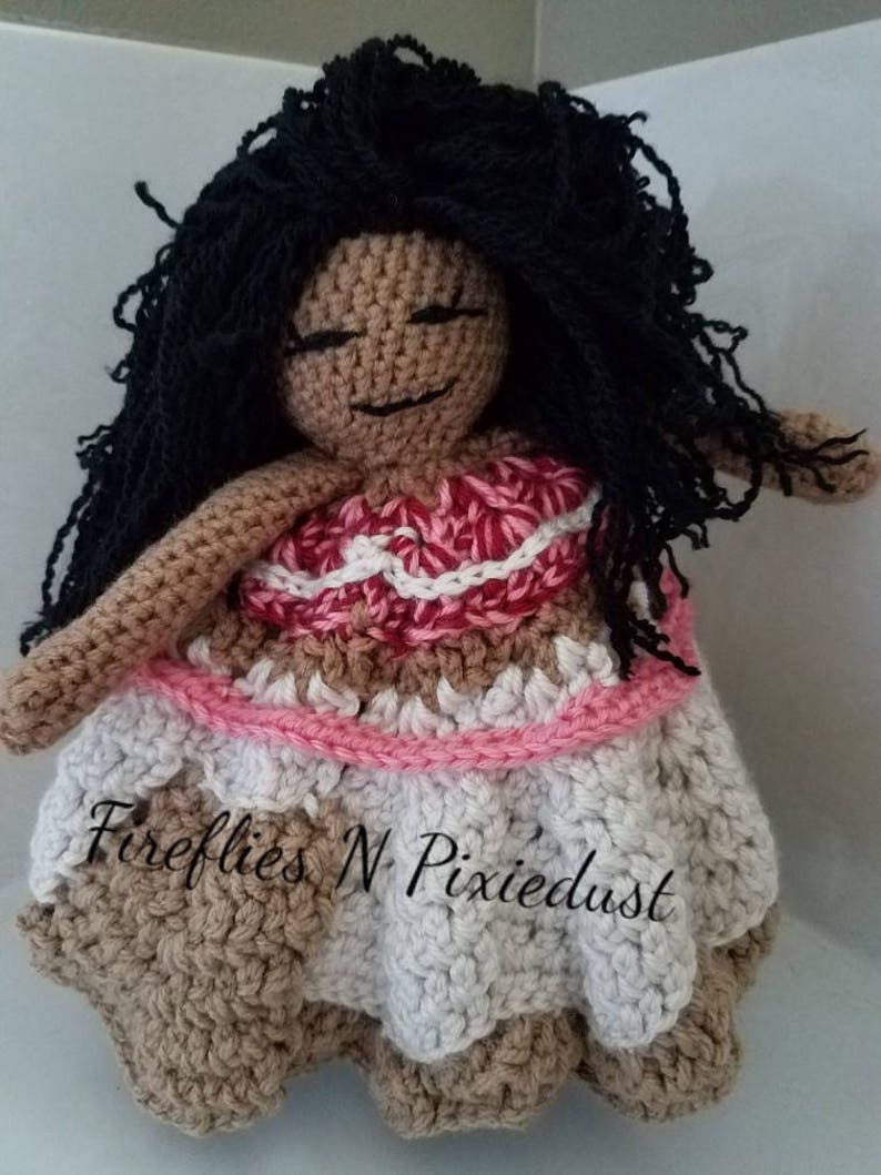 Puede incluir: Mu&ntilde;eca de crochet con piel marr&oacute;n, cabello negro y un vestido rosa y blanco. La mu&ntilde;eca lleva un vestido rosa y blanco con una falda marr&oacute;n. La mu&ntilde;eca sostiene una flor en su mano. La mu&ntilde;eca est&aacute; sentada sobre un fondo blanco. El texto "Fireflies N Pixiedust" es visible en la falda de la mu&ntilde;eca.