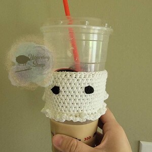 May include: A white crocheted coffee cup cozy in the shape of a ghost with two black eyes. The cozy is wrapped around a clear plastic cup with a red straw. The cup is filled with a light brown beverage.
