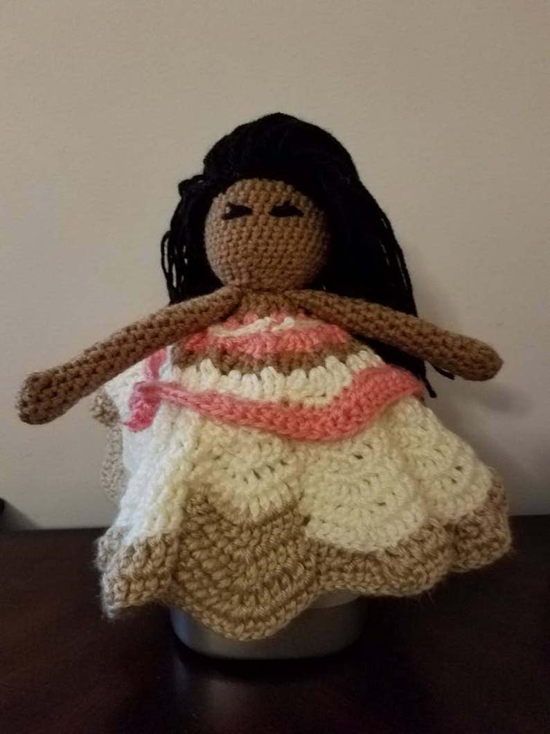 Puede incluir: Mu&ntilde;eca de crochet con piel marr&oacute;n, cabello negro y un vestido rosa y blanco. La mu&ntilde;eca lleva un vestido de crochet con un patr&oacute;n rosa y blanco.