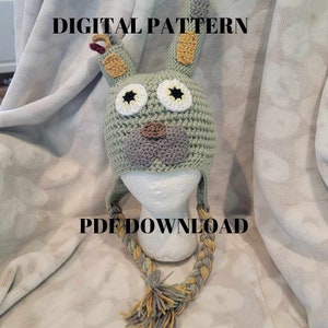 May include: A crochet pattern for a green bunny hat with a long braided tail. The hat has a gray nose, brown mouth, and large white eyes with black pupils. The pattern is available as a PDF download.