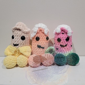 May include: Three whimsical, handmade crochet figures in shades of beige, peach, and pink, each with a unique design. The figures have black button eyes and embroidered smiles. The figures are displayed on a white surface.
