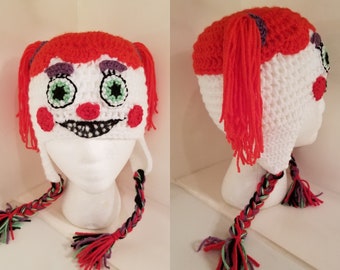 Puede incluir: Un gorro blanco de crochet con una cara roja y blanca, coletas rojas y un cordón trenzado multicolor. El gorro está en una cabeza de maniquí blanca.