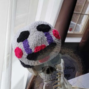 May include: A white crocheted plush doll head with black, purple, and pink accents. The doll has black eye patches, purple and pink teardrops, and a black mouth. The doll is part of a Halloween or horror-themed craft.