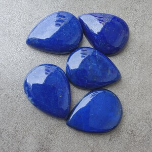 May include: Five polished lapis lazuli gemstones shaped like teardrops. Each stone features a deep royal blue color with subtle, natural golden pyrite flecks scattered across the smooth, reflective surfaces of these cabochon-style stones.