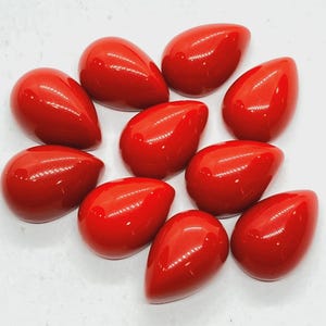 May include: A collection of nine smooth, teardrop-shaped cabochons in a vibrant, glossy red finish. Each piece features a rounded, domed top and a flat base, reflecting light with a polished sheen. The items are arranged in a loose cluster.