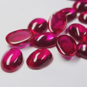 Può includere: Una collezione di pietre preziose cabochon ovali sfuse in un colore rosso magenta profondo e vibrante. Ogni pietra presenta una superficie liscia, lucida e arrotondata che riflette la luce, creando riflessi luminosi sui loro corpi traslucidi.