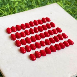 May include: A collection of small, smooth, oval-shaped red cabochons arranged in a grid pattern. Each piece features a glossy, solid red finish with a rounded top surface, uniform in size and shape, displayed on a flat, off-white square surface.